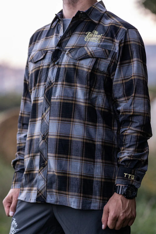 A man wearing a dark gray and black plaid button-up shirt with gold accents, featuring a 'Tactical Gear' logo on the left chest and 'TTG' on the right cuff, set against a blurred outdoor background.