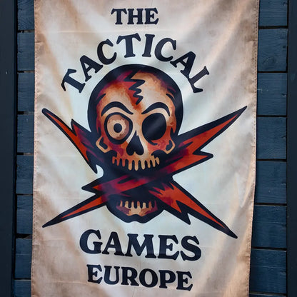 A vertical fabric banner, flag with a skull and lightning bolt graphic, reading 'THE TACTICAL GAMES EUROPE' in black text on a cream background, hanging against dark blue wooden siding.
